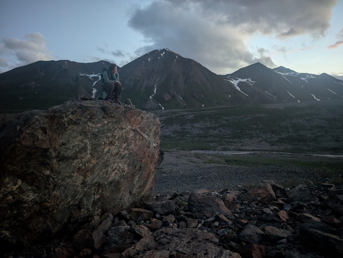 In low twilight, a person sits hunched on top of a large boulder with knees drawn up, gazing out across a broad gravel-braided valley toward dark mountains streaked with old snow. Talus and scattered rock lie below the boulder, and a few clouds catch faint light at right in a dimming blue sky.