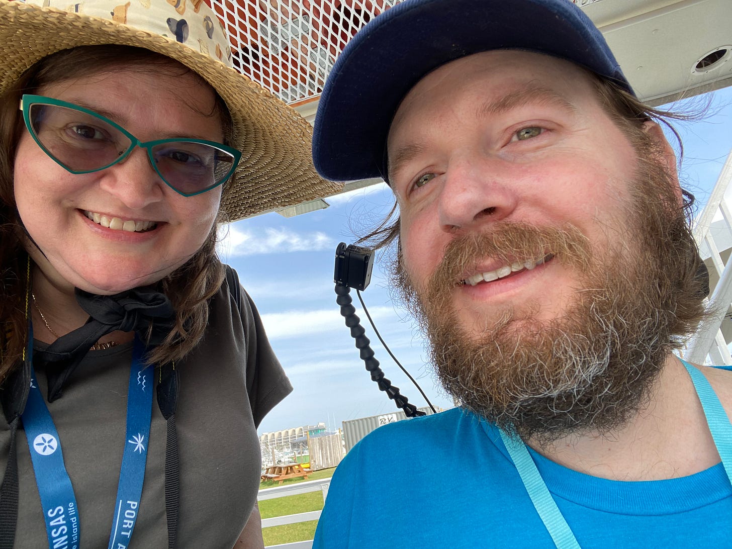 Eric and Nicole smile for a selfie on a bright afternoon. Eric wears a blue ball cap and a thick brown and gray beard. Nicole wears a straw shade hat and triangular green-rimmed glasses that recall a superhero’s mask. The words “Port Aransas” are printed on their lanyards.