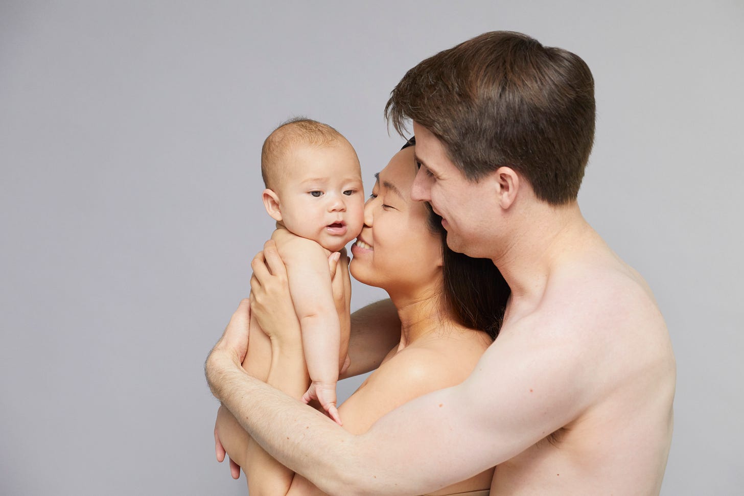 A young couple embraces their baby, all unclothed, in a warm and tender family portrait against a neutral gray background. A young couple embraces their baby, all unclothed, in a warm and tender family portrait against a neutral gray background.