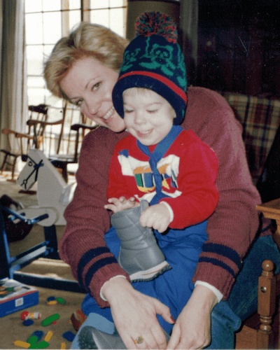 A woman smiling while playfully helping a young child put on their boots, set in a cozy indoor environment.