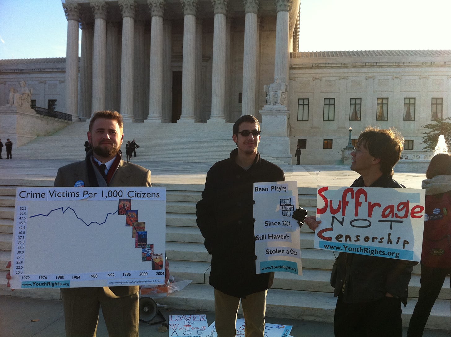 Three men standing in front of the U.S. Supreme Court building holding signs. The middle one states: "“Been playing Grand Theft Auto since 2004. Still haven’t stolen a car.” Three men standing in front of the U.S. Supreme Court building holding signs. The middle one states: "“Been playing Grand Theft Auto since 2004. Still haven’t stolen a car.”