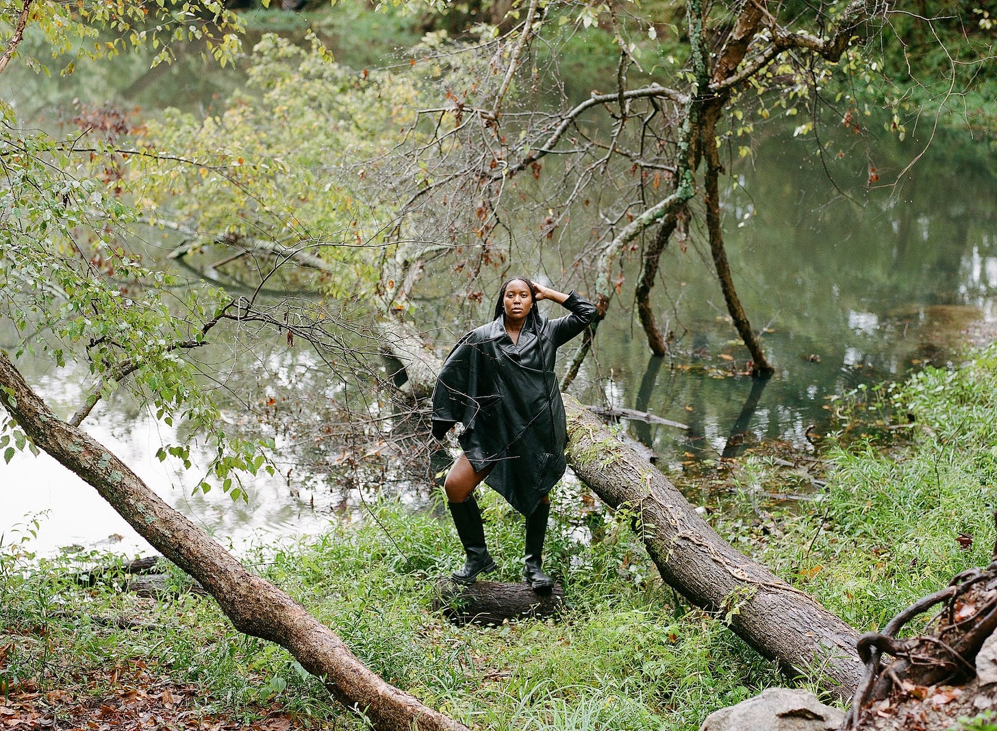 I am standing on a log at the edge of a swamp wearing knee high rubber boots and a reconstructed leather jacket from the Cykofa collection. Box braids greet my hips and my hands greet my waist and head and in a moment of stillness, I gaze directly at the camera. I am standing on a log at the edge of a swamp wearing knee high rubber boots and a reconstructed leather jacket from the Cykofa collection. Box braids greet my hips and my hands greet my waist and head and in a moment of stillness, I gaze directly at the camera.