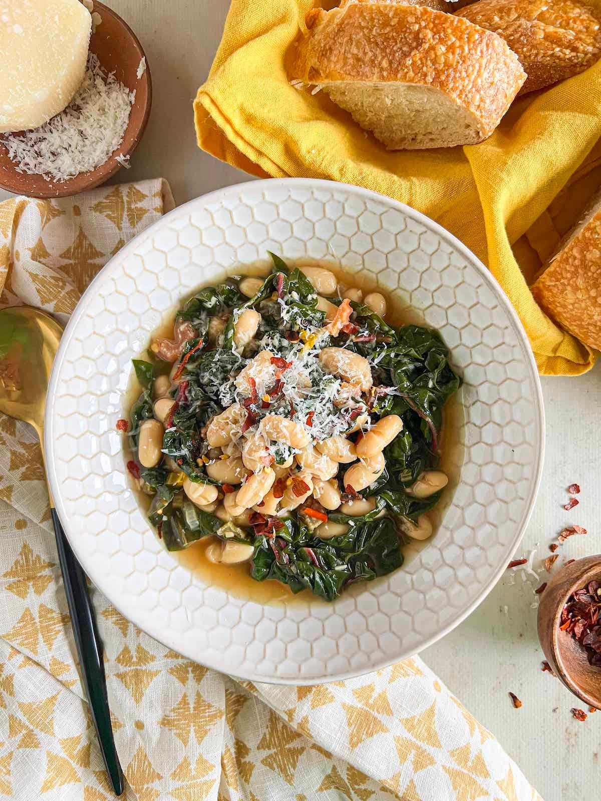 Beans and greens topped with parmesan in a white patterned bowl surrounded by a bread basket, yellow napkins, hunk of parmesan and small bowl of red pepper flakes.