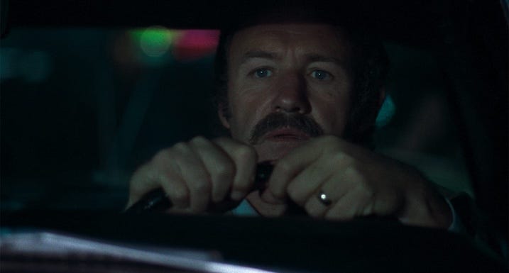 Gene Hackman grips a steering wheel tightly in a night shot, his expression tense and eyes focused ahead.