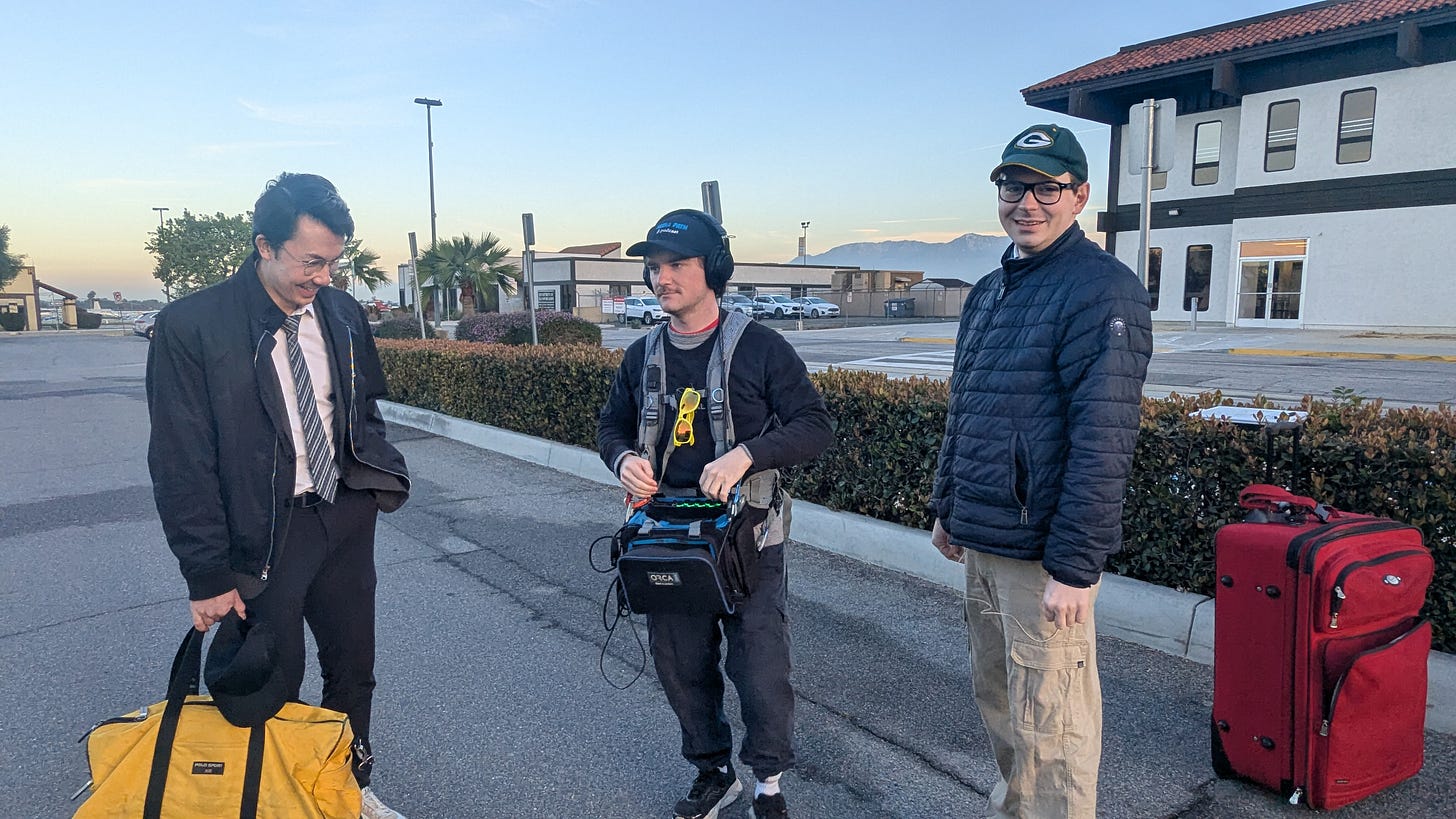 Three members of the production stand in a parking lot outside a small airport building at dusk. One wears a suit and holds a yellow duffel bag, another carries sound recording equipment with headphones, and a third stands beside a red suitcase. Mountains and a low building are visible in the background.