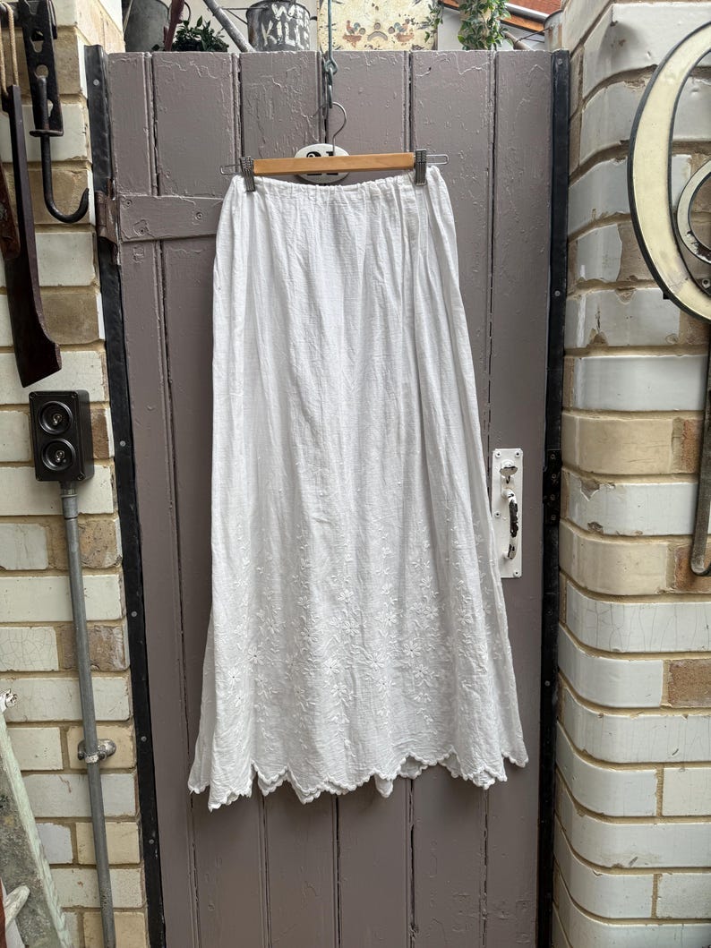 May include: A white, floor-length skirt with an elastic waistband and intricate floral embroidery. The skirt has a scalloped hemline and is hanging on a wooden hanger against a weathered wooden door.
