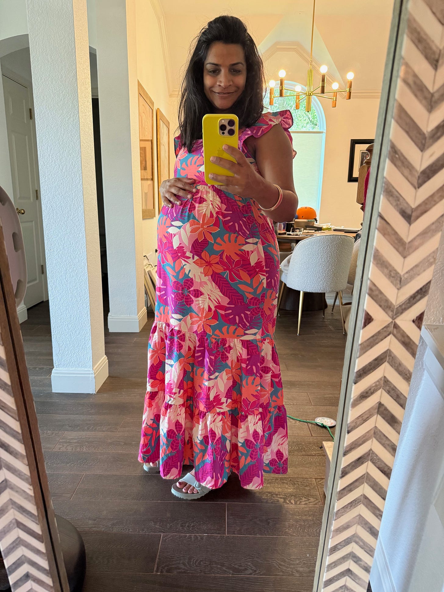 A woman wearing a pink floral maxi dress takes a selfie in a mirror. She is pregnant and has her right hand on her belly.