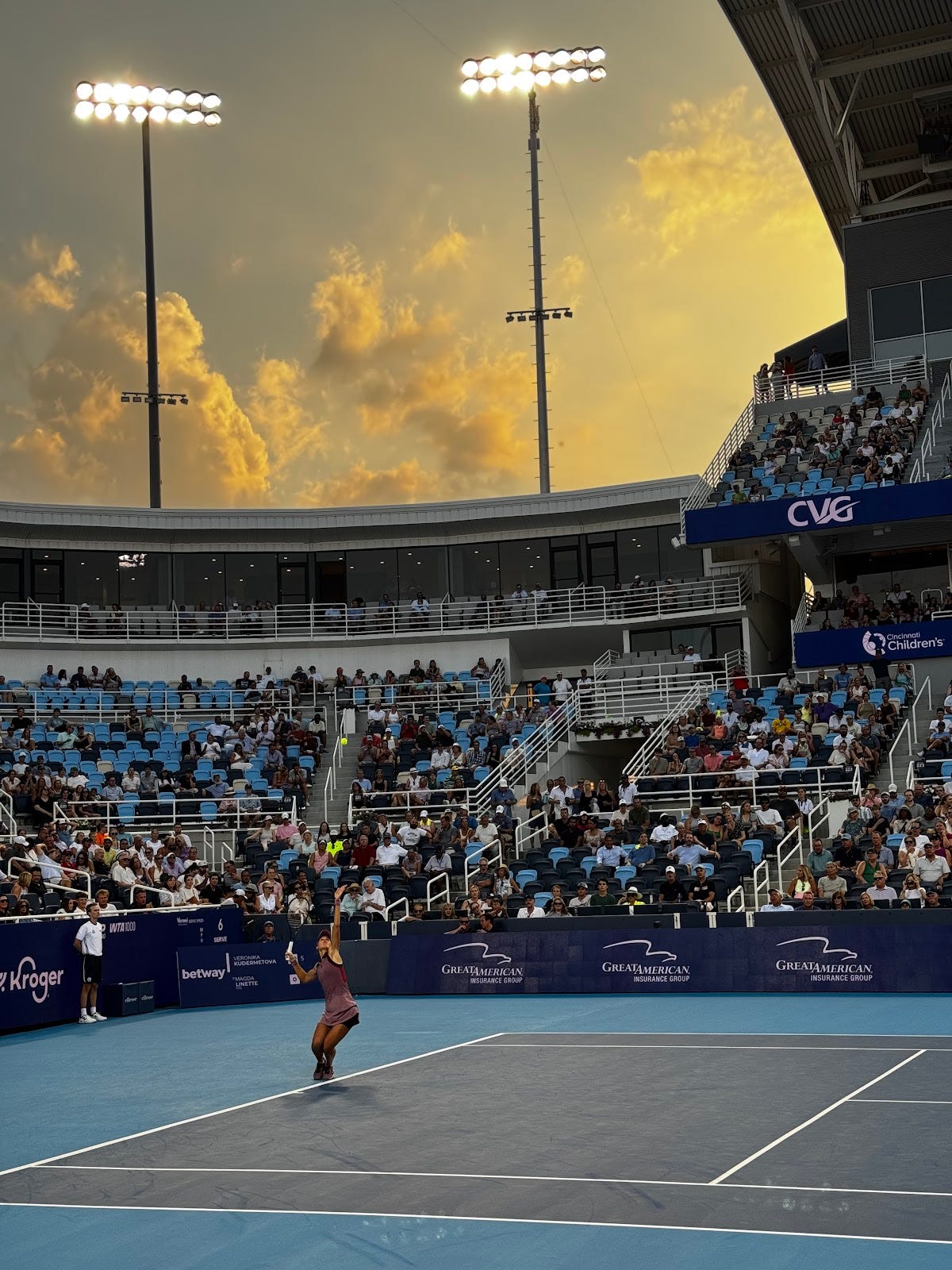 Dramatic golden sunset sky over Cincinnati Open tennis stadium during evening match, professional tennis player competing on blue hard court with CVG and Great American Insurance Group signag