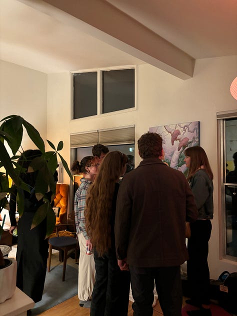 Photo 1: People gathering in a living room with vaulted ceilings. Photo 2: A painting and 3 drawings hung on a wall. Photo 3: A surrealist painting of amorphous creatures hanging from a tree. Photo 4: People gathered around the surrealist painting. Photo 5: A collage with cartoon images of a trail to the ocean and a butterfly, and bold text reading: traces of a path, losing sight of the sea. Photo 6: People and a red dog gathered around a man playing guitar. Photo 7: Ink and pastel drawings of two circles meeting. Photo 8: a pile of shoes at a door. Photo 9: Silhouettes of people through tall windows from outside at night.