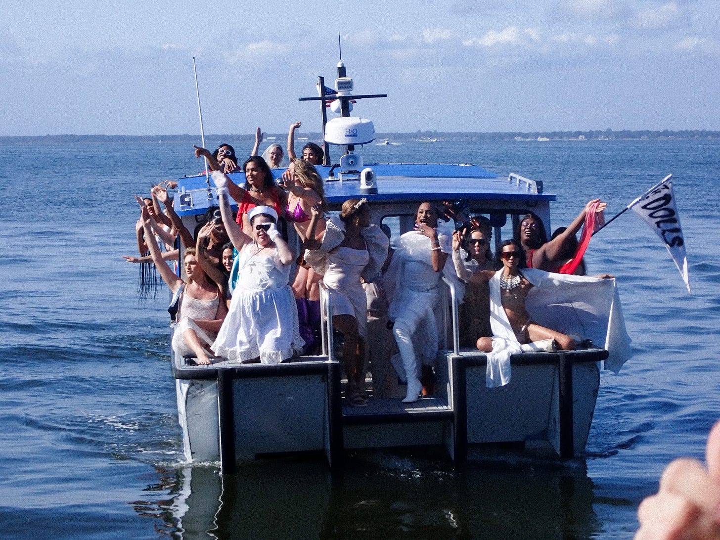 A group of people on a boat wearing various white outfits, celebrating with raised arms and big smiles. A flag reading "Dolls" flutters at the side of the boat. The background features calm blue water and a clear sky.