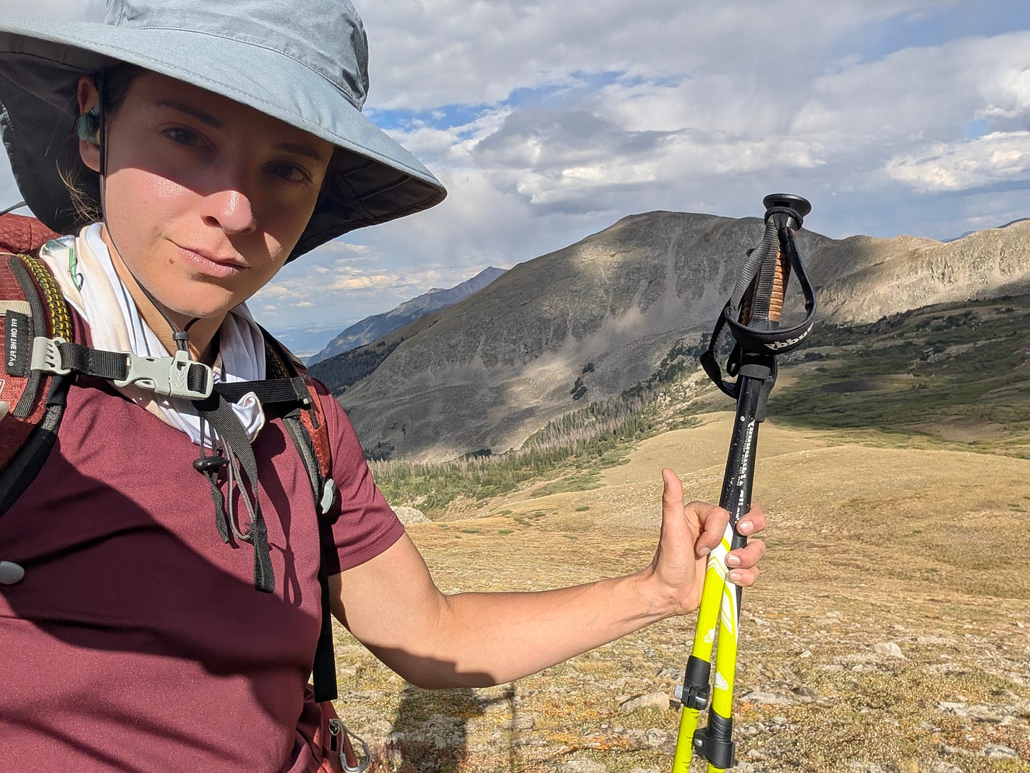 Selfie on trail in a sun hat holding trekking poles, with a broad mountain ridge and valley behind. Selfie on trail in a sun hat holding trekking poles, with a broad mountain ridge and valley behind.