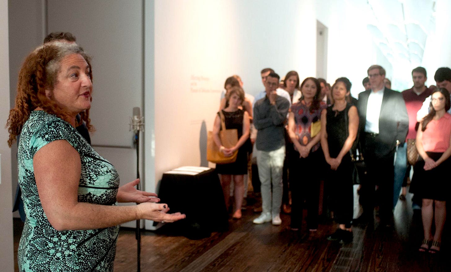 Claudia in an aqua blue and black dress speaks to a group of people who listen intently
