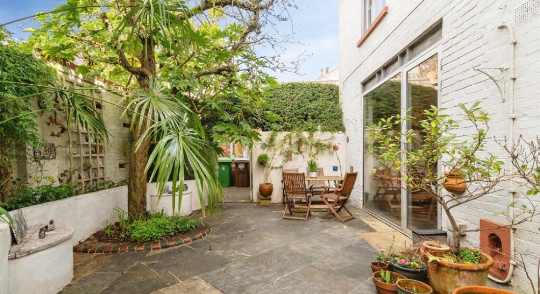 A house exterior with trees and shrubbery, and a grey patio A house exterior with trees and shrubbery, and a grey patio