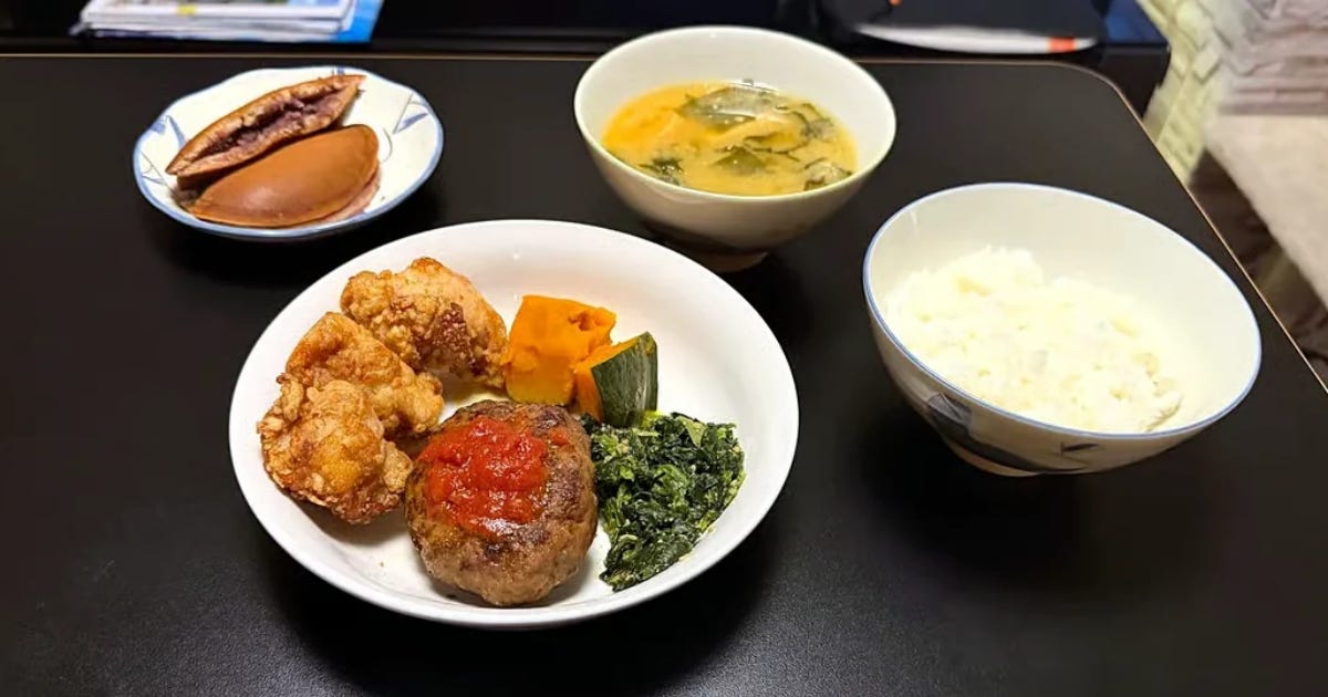 A Japanese-style dinner on a dark table. The main plate holds a hamburger steak topped with tomato sauce, three pieces of karaage fried chicken, simmered pumpkin and a small mound of spinach. Beside it are a bowl of white rice and a bowl of miso soup with wakame seaweed. A small plate at the back holds two dorayaki pancakes, one cut open to show the sweet red bean filling inside. A Japanese-style dinner on a dark table. The main plate holds a hamburger steak topped with tomato sauce, three pieces of karaage fried chicken, simmered pumpkin and a small mound of spinach. Beside it are a bowl of white rice and a bowl of miso soup with wakame seaweed. A small plate at the back holds two dorayaki pancakes, one cut open to show the sweet red bean filling inside.