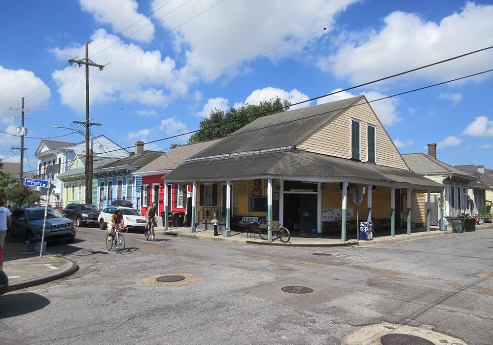 A corner convenience store in the Bywater neighborhood in New Orleans, a city with a long history of corner shops in predominantly residential areas (Zoning Practice December 2025) A single-story, siding-clad wooden commercial structure, with a hipped-roof overhang shading the entrance of a store, at the corner of two residential streets lined by modest shotgun homes.