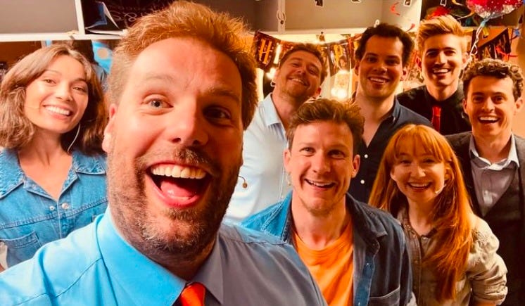 8 performers smile broadly for a selfie. They are mostly wearing shades of blue with some orange accessories