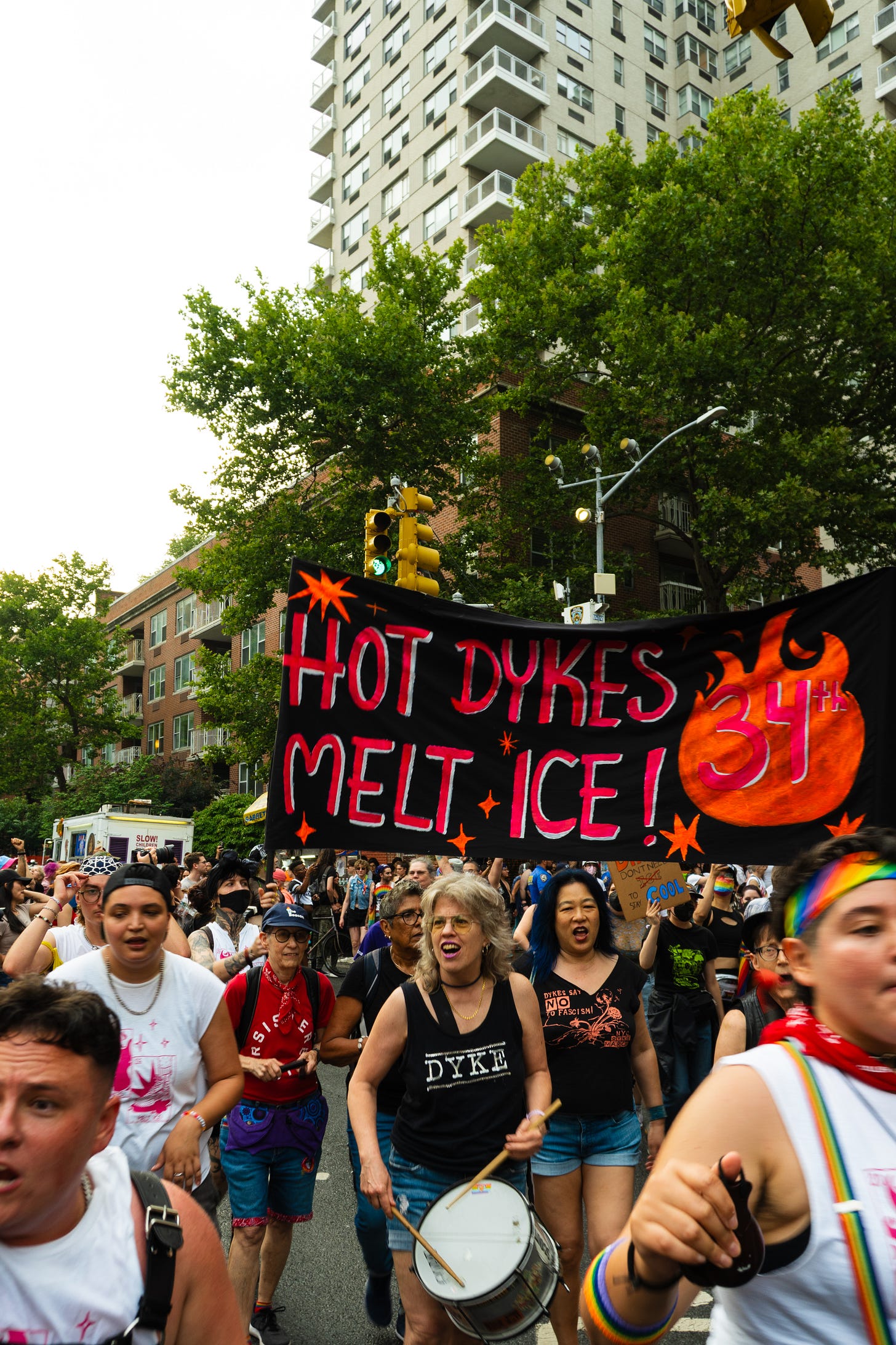 A drummer with gray hair, glasses, and a shirt that reads "dyke" is banging on their drum in front of the march banner that reads" Hot Dykes Melt Ice 34th Annual NYC Dyke March"