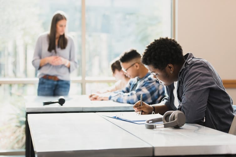 Three students are seen in a row leaning over papers on a table as a woman stands nearby them. Three students are seen in a row leaning over papers on a table as a woman stands nearby them.