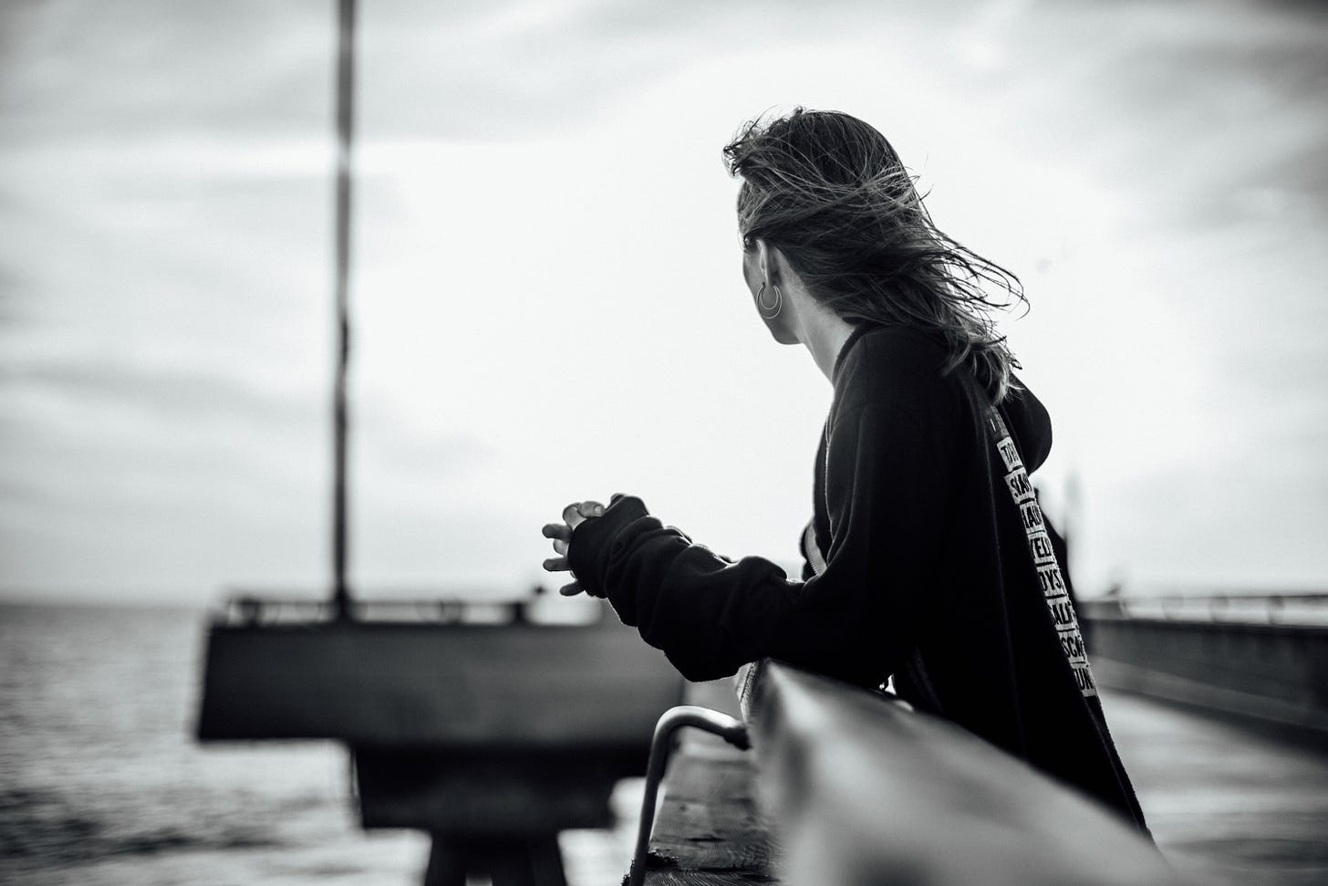 black and white photo of Kari on the pier at Venice Beach, wearing a black hoodie with white writing and gazing out at the ocean