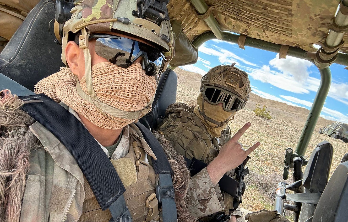2 young men wear camouflage uniforms and helmets, their faces are covered with sunglasses and scarves. Sitting in a small, military offroad vehicle