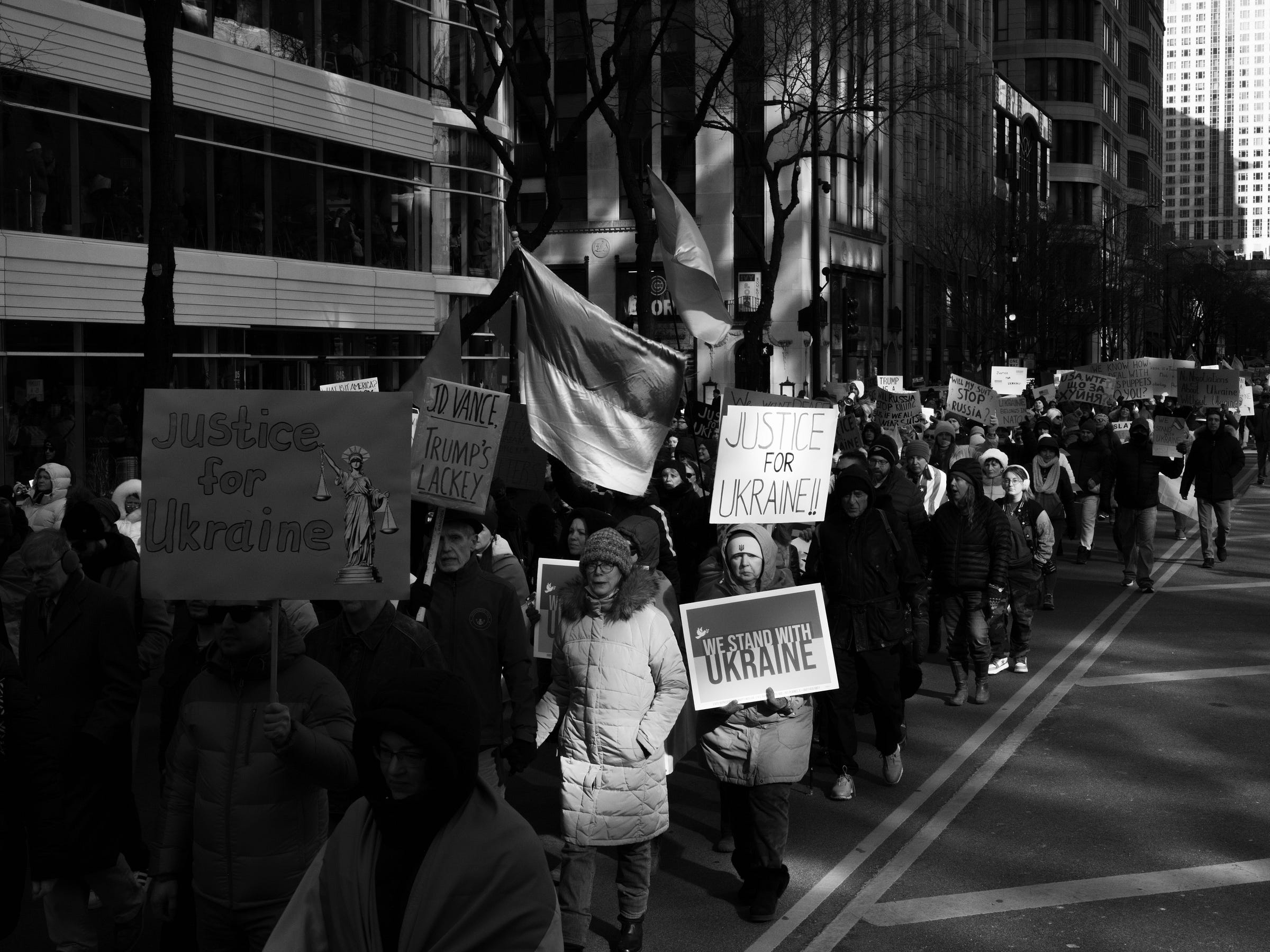 A black and white photograph captures a large crowd of protesters marching down a city street, holding signs and flags in support of Ukraine. Prominent signs read "Justice for Ukraine," "We Stand with Ukraine," and "JD Vance, Trump’s Lackey." Ukrainian flags wave among the demonstrators as they walk between tall buildings, their expressions determined and resolute. Sunlight and shadows highlight the movement of the march. A black and white photograph captures a large crowd of protesters marching down a city street, holding signs and flags in support of Ukraine. Prominent signs read "Justice for Ukraine," "We Stand with Ukraine," and "JD Vance, Trump’s Lackey." Ukrainian flags wave among the demonstrators as they walk between tall buildings, their expressions determined and resolute. Sunlight and shadows highlight the movement of the march.