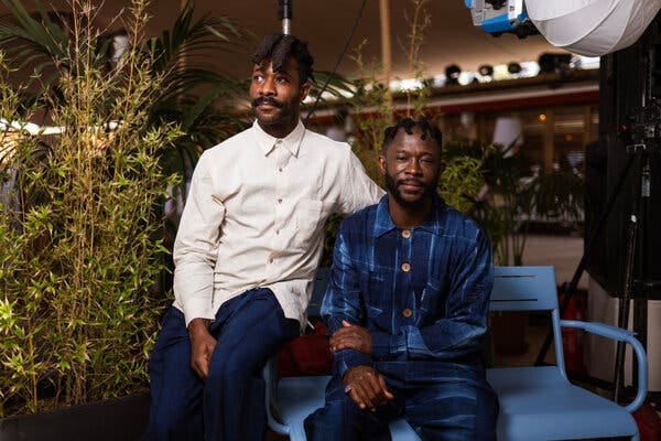 A man in an untucked white shirt sits on the arm of a blue chair next to a man in dark blue. A man in an untucked white shirt sits on the arm of a blue chair next to a man in dark blue.