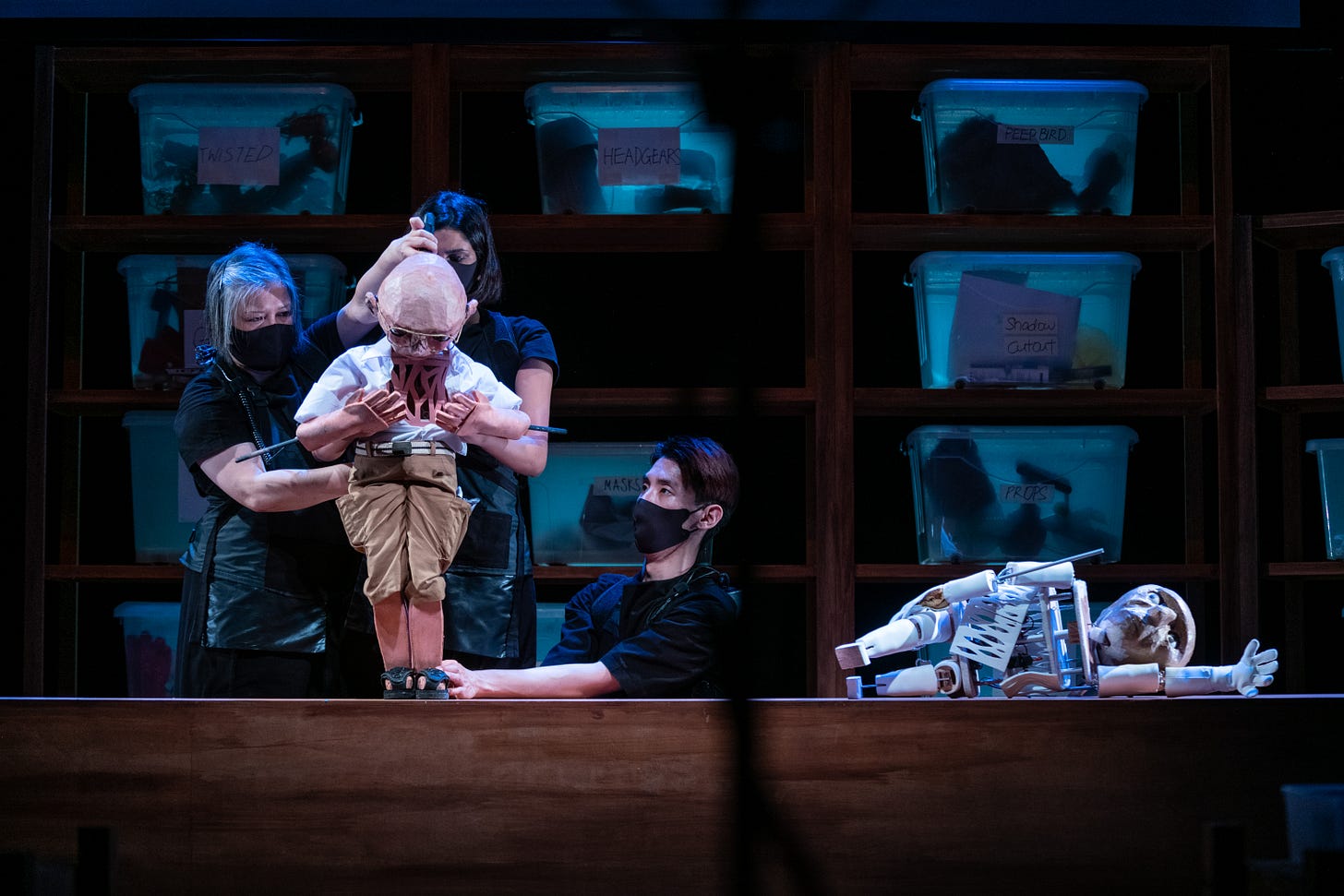 The animatronic puppet Chiam, supported by three puppeteers, unbuttons the front of his shirt to reveal the mechanics in his chest. Next to him, a similar-looking puppet lies lifeless on a workbench.
