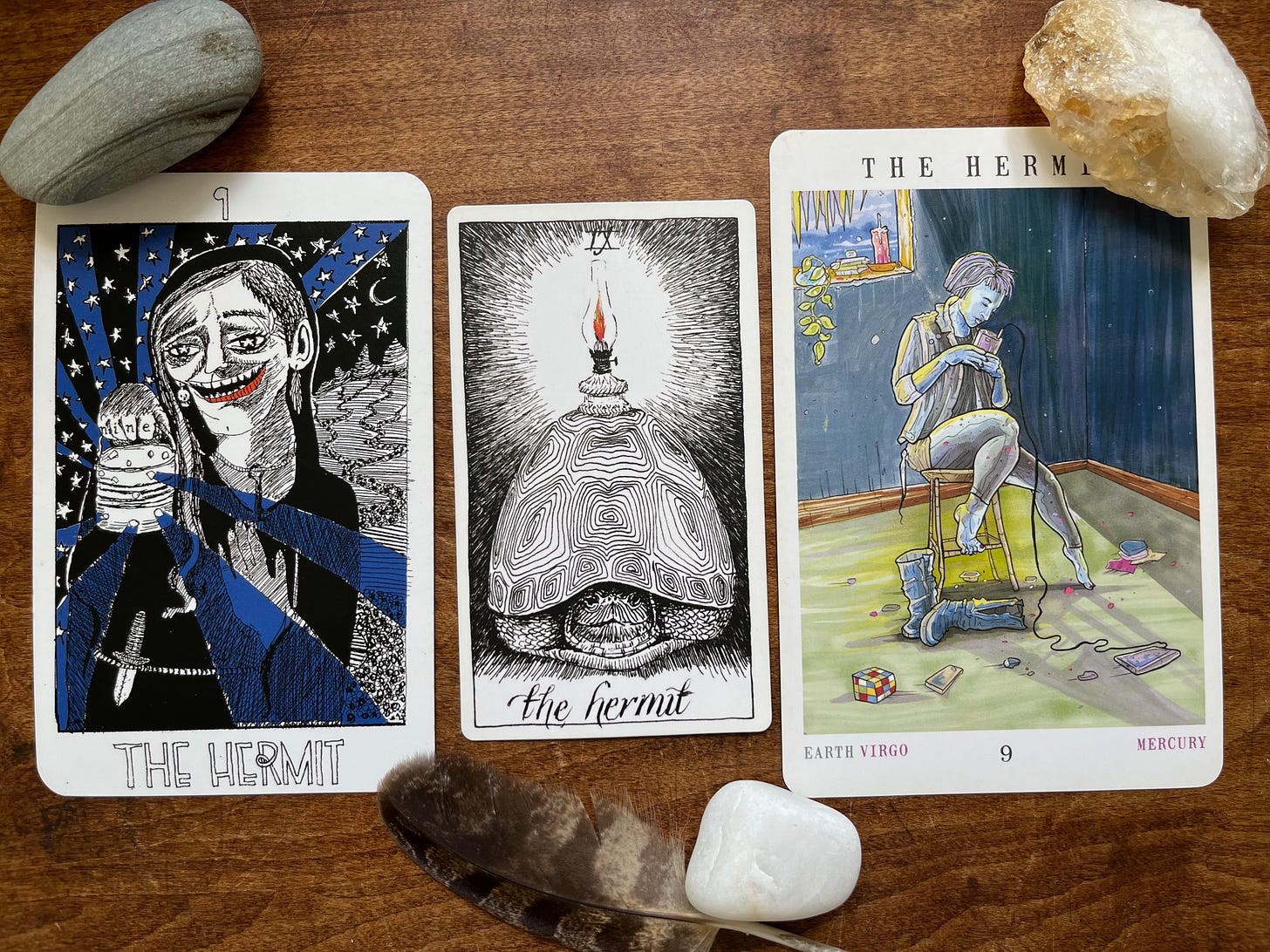 Three versions of the Hermit card, surrounded by three stones and a small feather. The left card, from the Collective Tarot, depicts a person walking under a starry sky in the wilderness, holding up a lamp with small holes that beam out light. They are smiling, they wear a small knife at their waist, and the hand holding the lamp has the word “nine” tattooed on their fingers. The middle card, from The Wild Unknown Tarot, is a black-and-white drawing of a tortoise looking out, with a small oil lamp with an orange flame balanced on their shell. The card on the right, from the Next World Tarot, shows a person alone in a mostly bare room, sitting on a stool, boots off, looking at their phone. There’s a Rubik's Cube on the floor and a candle, crystals, and a plant on the windowsill behind them. Three versions of the Hermit card, surrounded by three stones and a small feather. The left card, from the Collective Tarot, depicts a person walking under a starry sky in the wilderness, holding up a lamp with small holes that beam out light. They are smiling, they wear a small knife at their waist, and the hand holding the lamp has the word “nine” tattooed on their fingers. The middle card, from The Wild Unknown Tarot, is a black-and-white drawing of a tortoise looking out, with a small oil lamp with an orange flame balanced on their shell. The card on the right, from the Next World Tarot, shows a person alone in a mostly bare room, sitting on a stool, boots off, looking at their phone. There’s a Rubik's Cube on the floor and a candle, crystals, and a plant on the windowsill behind them.