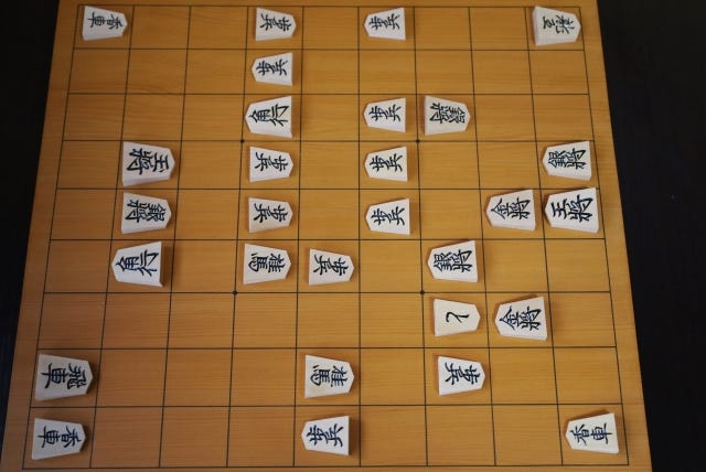 A shogi game in progress, photographed from above the board. The wooden board has a 9x9 grid, and there are many pentagonal wooden pieces, inscribed with black kanji characters to indicate the piece type. The pieces point to the right or the left, indicating which player has the piece. A shogi game in progress, photographed from above the board. The wooden board has a 9x9 grid, and there are many pentagonal wooden pieces, inscribed with black kanji characters to indicate the piece type. The pieces point to the right or the left, indicating which player has the piece.