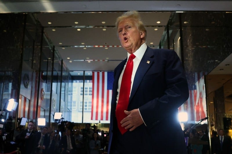 Trump is shown from below, mid-speech, wearing a navy blue suit and a thick red tie. An American flag is hanging behind him. Trump is shown from below, mid-speech, wearing a navy blue suit and a thick red tie. An American flag is hanging behind him.