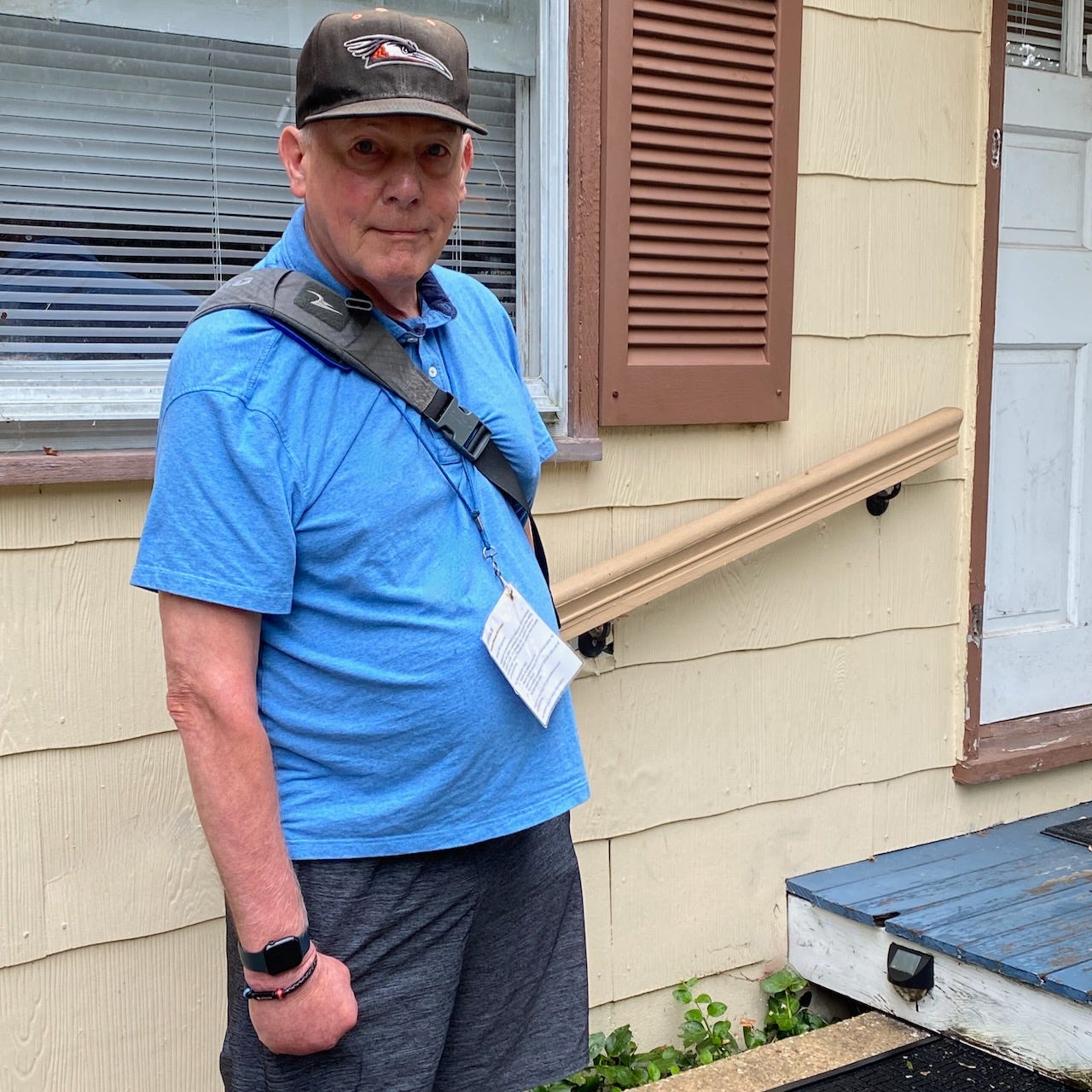 A man in a baseball hat standing outside a house, wearing a laminated identification card on a lanyard around his neck.