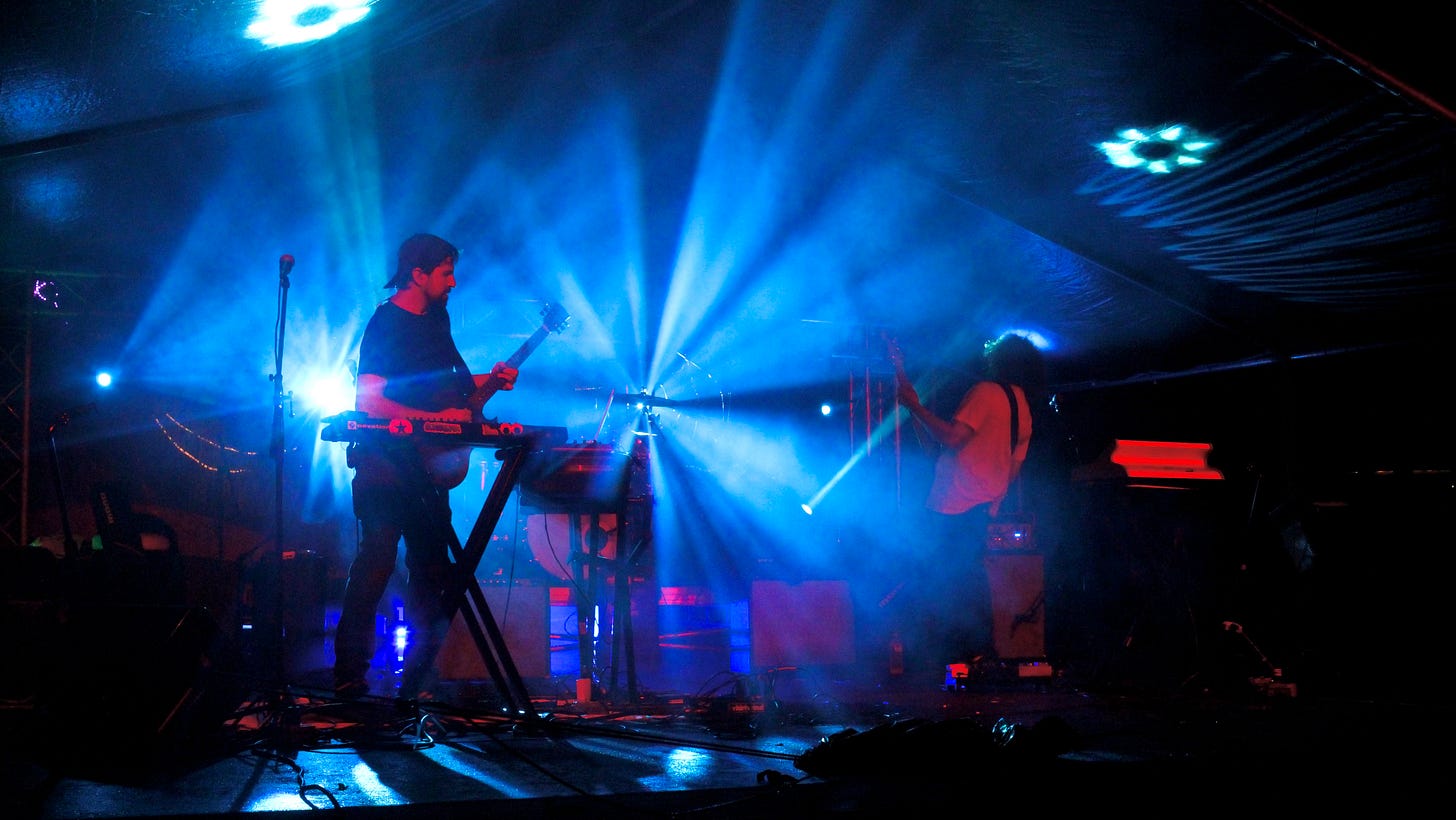 Professional concert photo of SunSquabi performing on the MoonGroove main stage at night, band members lit by colorful stage lighting with the crowd visible in the foreground. Professional concert photo of SunSquabi performing on the MoonGroove main stage at night, band members lit by colorful stage lighting with the crowd visible in the foreground.