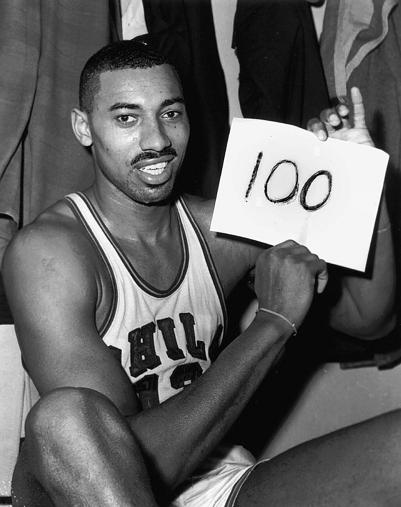 Wilt Chamberlain, an African American man, is shown sitting down in his Philadelphia Warriors jersey while holding up a piece of paper with the number 100 written on it. The photograph was taken directly after the game and is in black and white. Wilt Chamberlain, an African American man, is shown sitting down in his Philadelphia Warriors jersey while holding up a piece of paper with the number 100 written on it. The photograph was taken directly after the game and is in black and white.