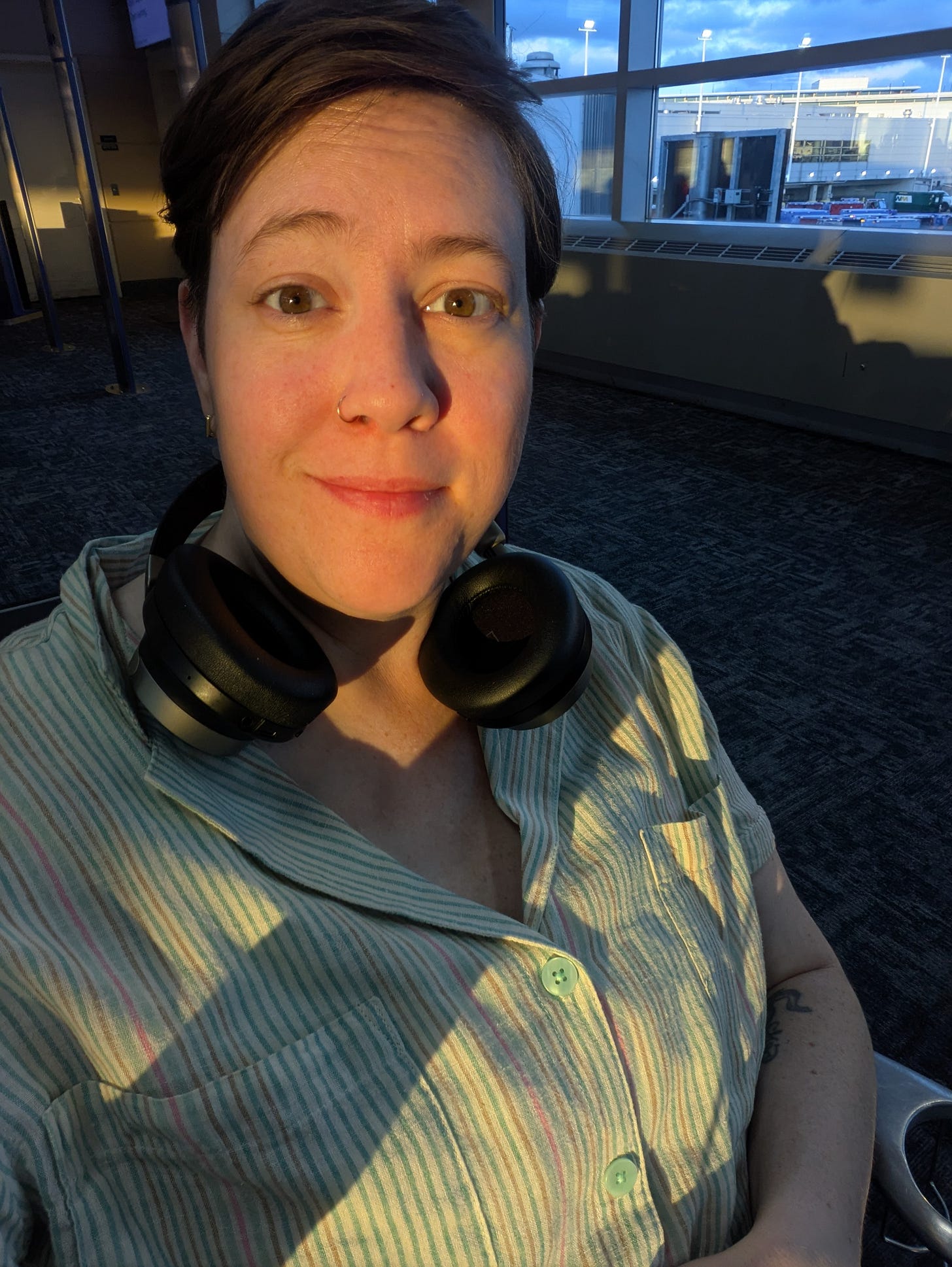 White woman sitting in an airport terminal with golden hour light falling across her face. She looks directly at the camera with a wry smile on her face. She is wearing a light green striped shirt and over-the-ear earphones around her neck.