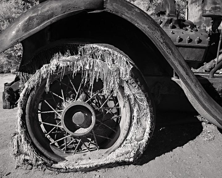 five photos that Kirk Marshall took. 1. B&W rolling sand dunes. 2. two metal dinosaur sculptures in a mock fight against a dark desert night. 3. B&W cactus along a road with the shadow of one across the road. 4. B&W an old destroyed tyre showing the decayed side walls while still attached to a rusted bumper. 5. A metal dragon sculpture created to look like it’s underneath the surface of the earth. A bright blue sky and some low lying hazy hills in the background.