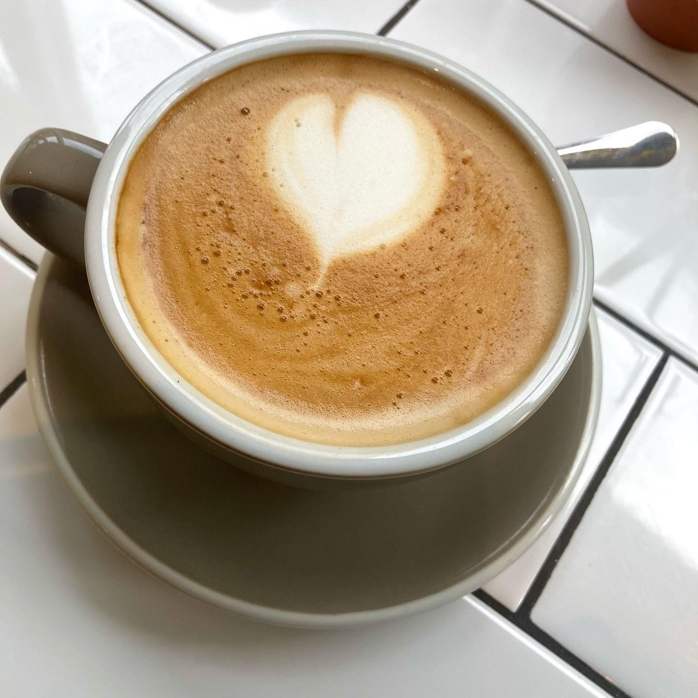 A coffee with a heart drawn in the foam on top of it, in a grey cup and saucer on a white tiled countertop in my favourite ice cream cafe in St Ives, Moomaid of Zennor.