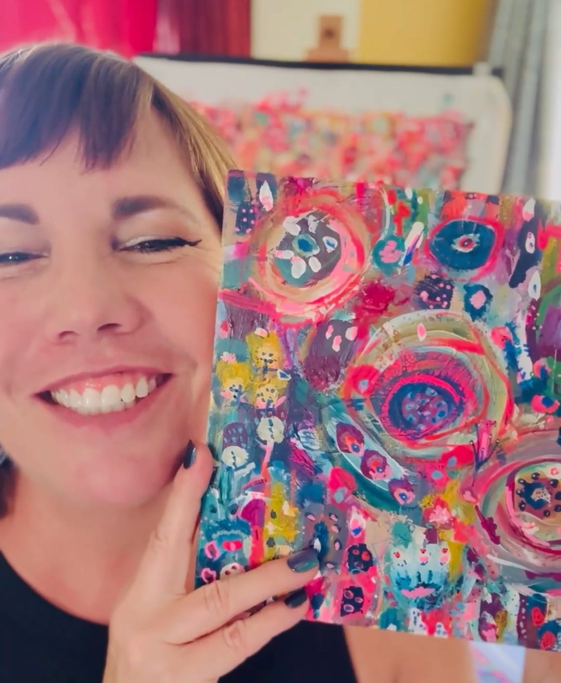 Artist Kelly Kingman smiling and holding one of her colorful abstract paintings featuring bright pinks, blues, and layered textures.