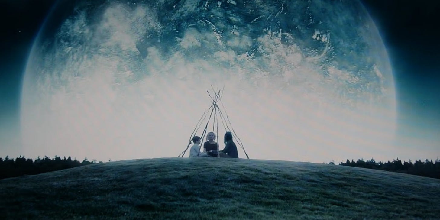 Three people, man, a woman, and a child, sit below a structure made of sticks in a clearing at night. Behind them is a massive planet, about to crash into the Earth.