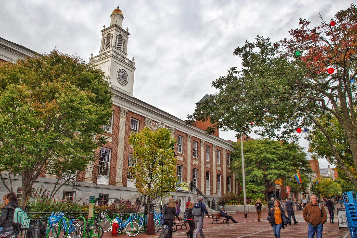 An exterior view of Burlington City Hall on a cloudy fall day, colorful leaves on the trees, people walking around on the sidewalk in front, bikes filling a bike rack. An exterior view of Burlington City Hall on a cloudy fall day, colorful leaves on the trees, people walking around on the sidewalk in front, bikes filling a bike rack.