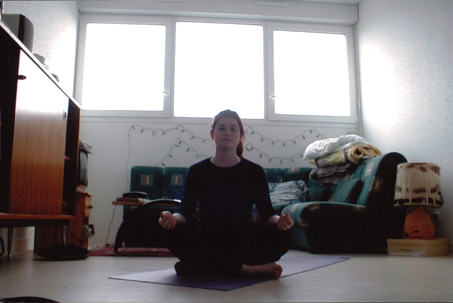 Charlotte sits in her apartment living room on a purple yoga mat with her legs folded and her wrists resting on her knees.