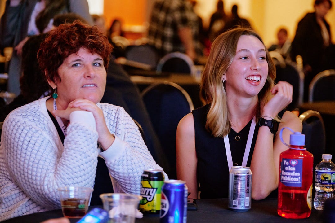 Two women seated side by side at a conference table, one leaning forward watching the stage intently, one laughing openly at something
