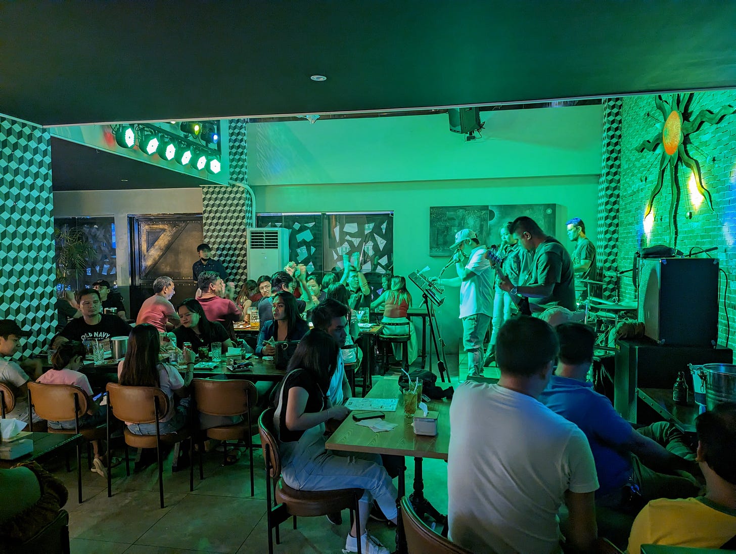 Photo inside a club in Manila. Tables filled with patrons are arranged around a stage where a singer and musicians are performing. The photo is tinted green from the green stage lighting.