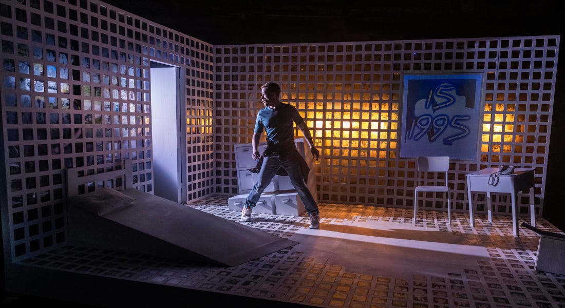 A man stands with legs wide, looking out a doorway, with a strong shadow behind him. The walls and floor are covered in a white grid pattern which is softly lit from behind. The figures 15 1995 are displayed on a panel on the wall. A man stands with legs wide, looking out a doorway, with a strong shadow behind him. The walls and floor are covered in a white grid pattern which is softly lit from behind. The figures 15 1995 are displayed on a panel on the wall.