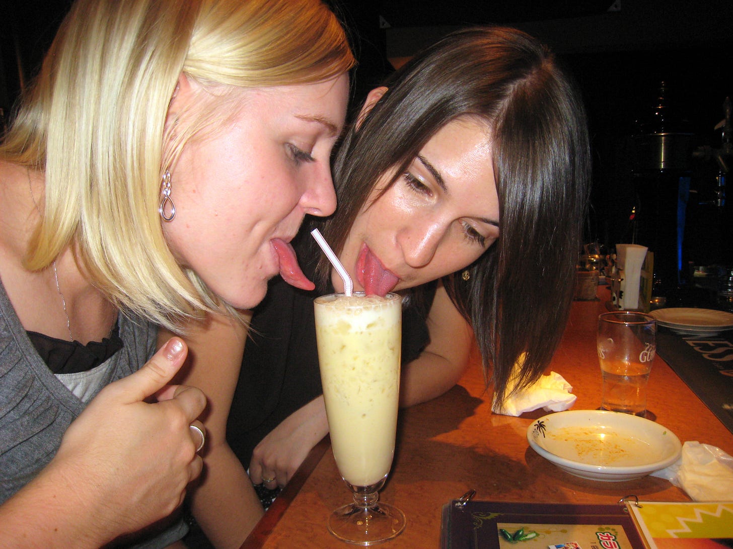 Two young women drink a piña colada cocktail from a single glass, their tongues sticking out Two young women drink a piña colada cocktail from a single glass, their tongues sticking out