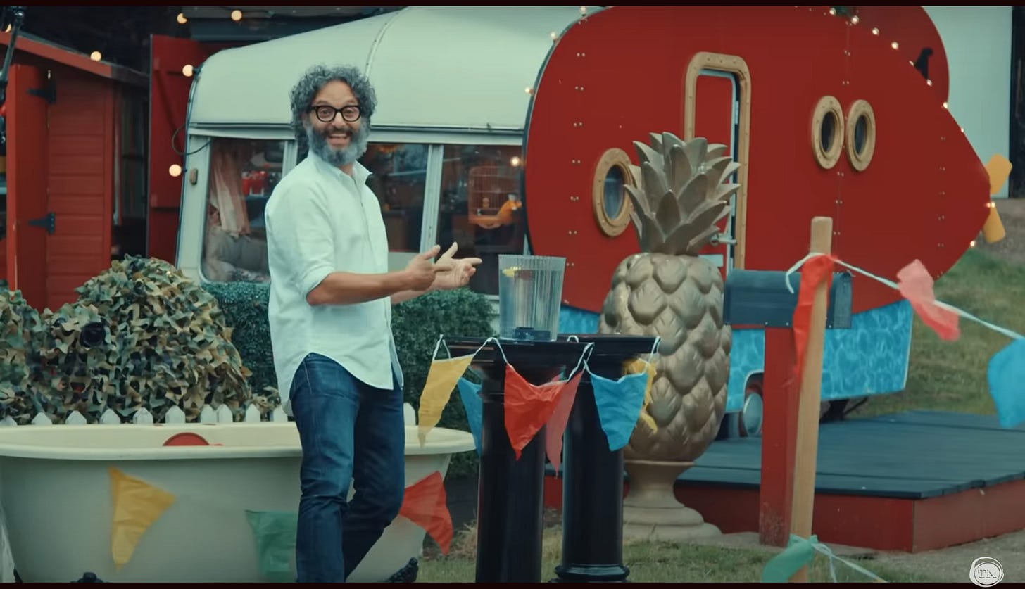 Jason Mantzoukas grins next to a vase not quite filled to the brim with water.