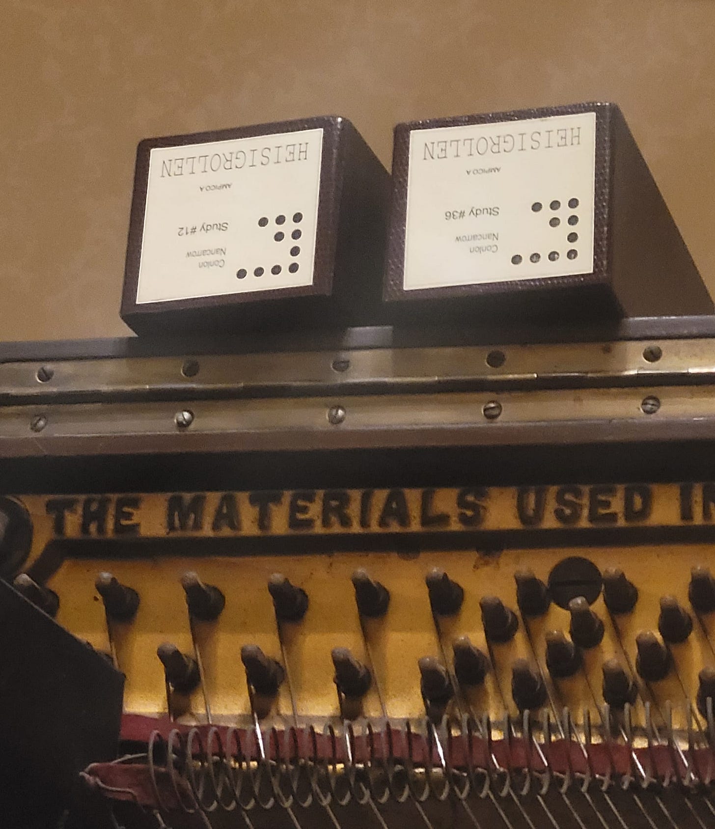 Close-up of two boxes containing the piano rolls for two studies by Conlon Nacarrow. They are resting upon the top of a player piano, on which can be read the embossed words 'The materials used'.
