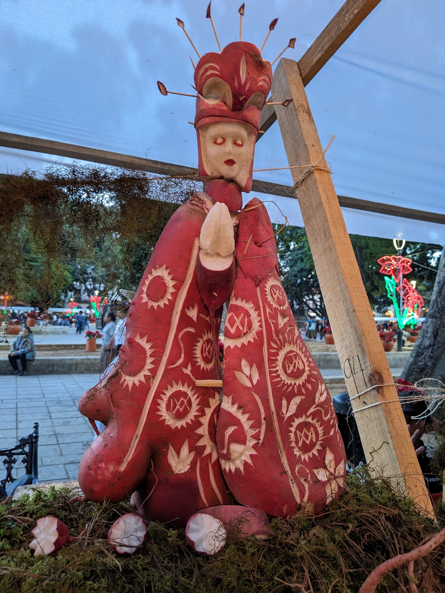 A woman with a white face and red lips is wearing a headdress and red dress and is roughly carved out of red and white radishes. 
