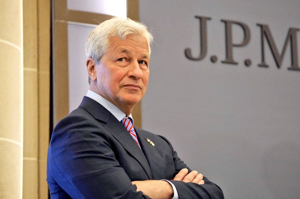 A man with white hair wearing a dark blue suit red tie and white shirt stands with arms crossed smiling slightly facing the camera. He has a serious expression and is positioned in front of a light stone wall with large metallic letters spelling JPM. The background includes architectural elements like columns suggesting a corporate headquarters setting.