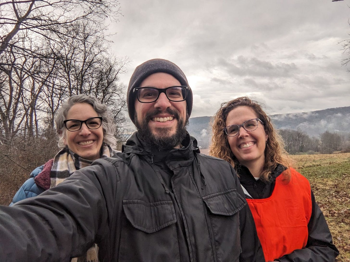 Three people standing in a field under a gray, cloudy sky. On the left in the back is me, with short wavy gray hair and glasses. In the center is Richard with a winter coat, hat, and glasses. He has a dark beard. On the right is Kathy, in a bright orange vest and glasses, with long wavy brown hair. We're all smiling.