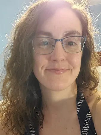 A headshot of a pale femme presenting person wearing glasses with long wavy brown hair and a smile with glossy lips. A headshot of a pale femme presenting person wearing glasses with long wavy brown hair and a smile with glossy lips.